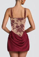 Rear view of a model wearing a short dress with a beige floral lace bodice and a draped burgundy skirt, set against a solid light gray background.