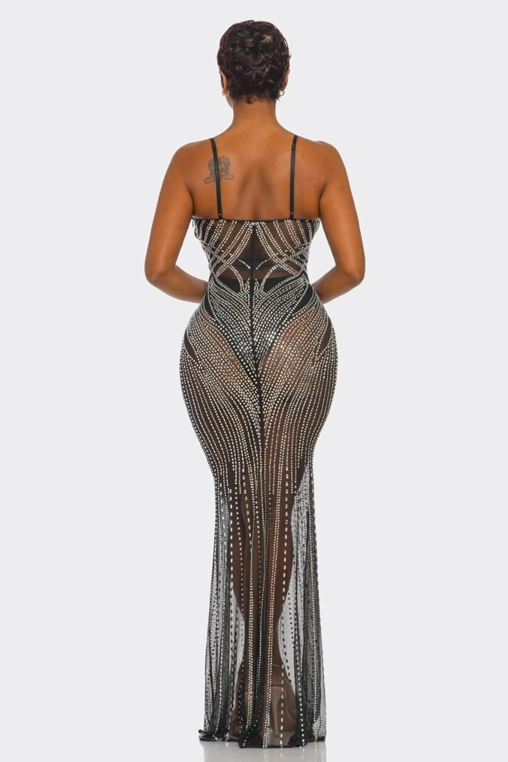 A woman viewed from the back wearing a sheer black mesh maxi dress heavily embellished with silver rhinestones, set against a plain light gray studio background.