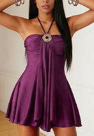 A woman wearing a purple halter mini dress with a ruched bodice, flared skirt, and a large jeweled circular pendant at the neckline.
