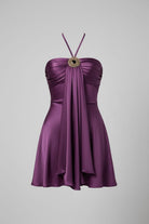 A woman wearing a strapless halter mini dress in deep purple with a large decorative circular brooch at the neckline, paired with silver bangles, set against a neutral indoor background.
