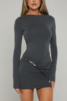 A woman wearing a dark gray long-sleeve bodycon mini dress with a twisted detail at the waist, standing in front of a white door with green garden foliage visible on the right.