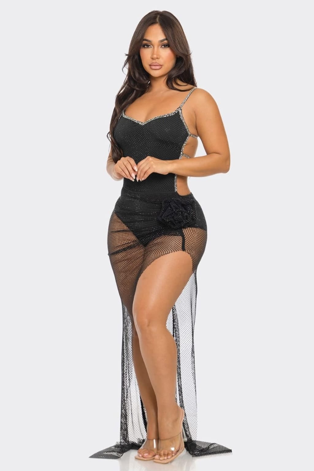 A woman modeling a black sheer mesh maxi dress with a sweetheart neckline, thin rhinestone-embellished straps, side cutouts, a floral waist detail, and a long trailing mesh overlay, photographed against a light gray studio background.