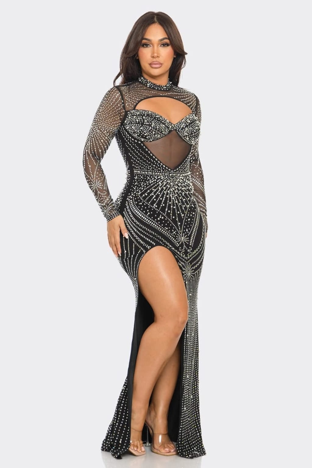 A woman modeling a long-sleeve, floor-length black gown covered in silver rhinestone and bead embellishments, featuring a high slit and sheer mesh details, set against a plain light gray background.