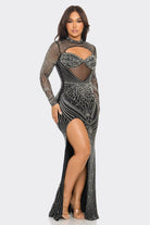 A woman modeling a long-sleeve, floor-length black gown covered in silver rhinestone and bead embellishments, featuring a high slit and sheer mesh details, set against a plain light gray background.