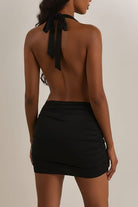 A woman wearing a black halter-neck mini dress with a wrap-style crossover bodice, side cutouts, and gathered draping details, photographed against a plain light beige studio background.