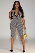 A woman modeling a black and white horizontally striped one-piece jumpsuit with a deep V-neckline and black trim, paired with black pointed-toe heels and a small gold handbag, standing outdoors at night.